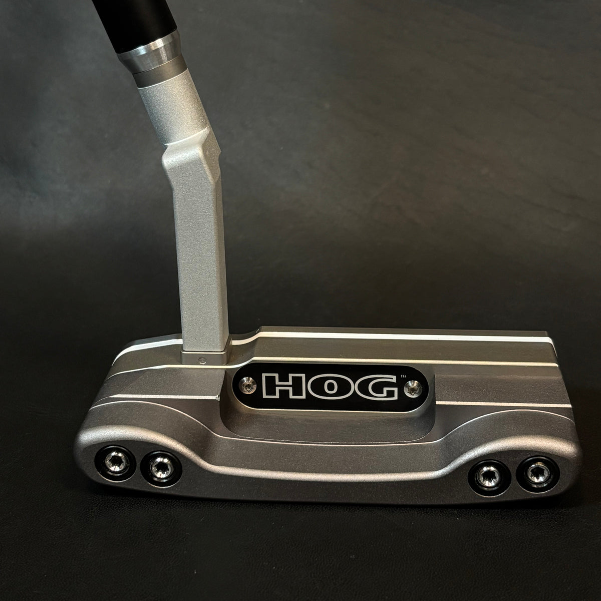 A close-up of a HOG golf putter head with a silver metallic body, black and silver handle, and a black logo plate on a dark gray background.