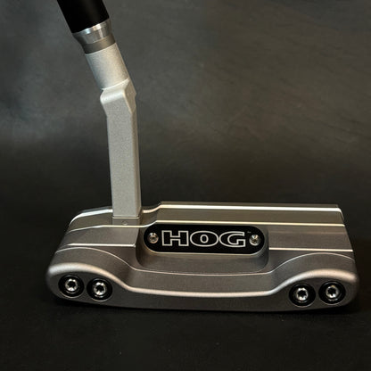 A close-up of a HOG golf putter head with a silver metallic body, black and silver handle, and a black logo plate on a dark gray background.