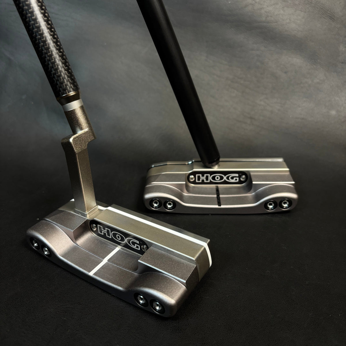 Two Hox golf putters with metallic silver and black heads and black shafts, displayed on a dark textured background.