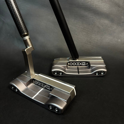 Two Hox golf putters with metallic silver and black heads and black shafts, displayed on a dark textured background.